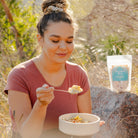 Woman eating outdoors with a bowl and spoon, holding a package of food.